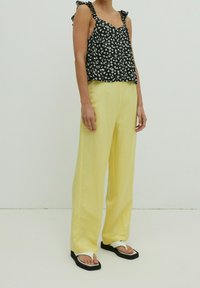 Top sin mangas negro con estampado floral y tirantes con volantes, combinado con pantalones amarillos holgados y sandalias negras con detalles blancos. Diseñado para un ajuste relajado.