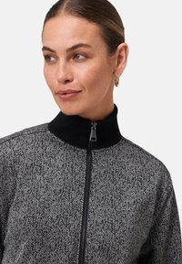 Femme avec les cheveux attachés portant un pull zippé noir et gris à motif avec un col haut et des boucles d'oreilles en or, regardant vers la gauche.