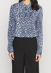 Chemise à imprimé léopard en bleu clair et marine, manches longues, col boutonné, texture soyeuse, associée à un pantalon noir taille haute.