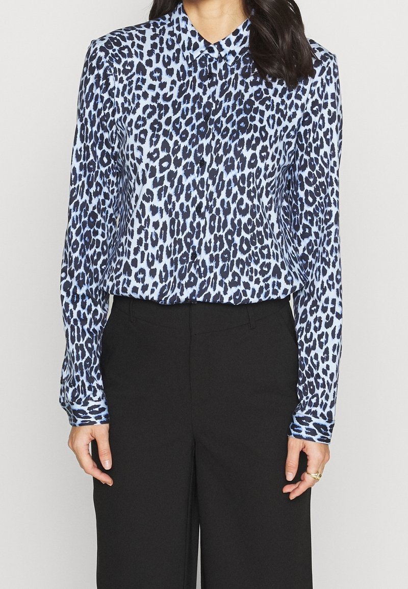 Chemise à imprimé léopard en bleu clair et marine, manches longues, col boutonné, texture soyeuse, associée à un pantalon noir taille haute.