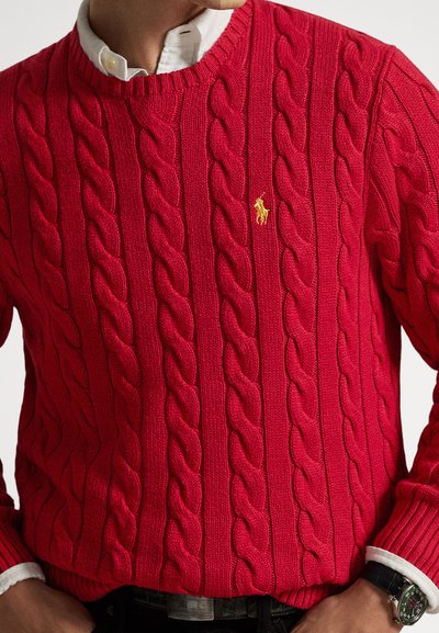 Άνδρας με κόκκινο πουλόβερ πλεκτό με καλώδια πάνω από λευκό πουκάμισο με γιακά, με τα χέρια στις τσέπες και ένα μαύρο ρολόι χειρός ορατό στον δεξί καρπό.