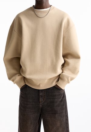 Homme portant un sweat-shirt beige oversize et un jean délavé foncé, les mains dans les poches, cadré du cou jusqu'à mi-cuisse.