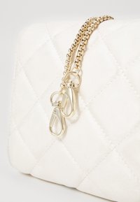 Bílá prošívaná taška vyrobená z měkkého materiálu, s zlatým popruhem na rameno a kováním na zavírání, s výrazným prošívaným vzorem.