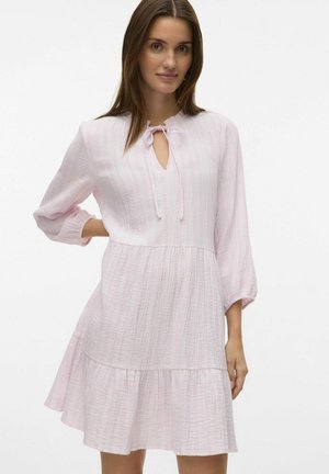 Robe en coton rose clair avec un tissu texturé, manches longues et un décolleté à lien en trou de serrure. Elle présente une jupe évasée avec un ourlet volants.