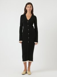 Γυναίκα που φοράει μαύρο μίντι φόρεμα με ριμπ ύφανση, κουμπιά σε σχήμα καρδιάς και μπεζ βολάν στις μανσέτες και το τελείωμα, συνδυασμένο με μπεζ παπούτσια.