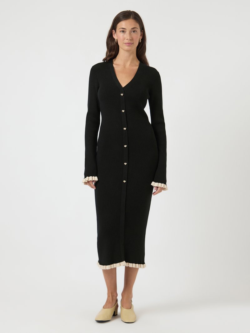 Γυναίκα που φοράει μαύρο μίντι φόρεμα με ριμπ ύφανση, κουμπιά σε σχήμα καρδιάς και μπεζ βολάν στις μανσέτες και το τελείωμα, συνδυασμένο με μπεζ παπούτσια.