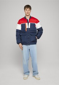 Prešívaná bomber bunda s tmavomodrým telom, červenými a bielymi panelmi, oranžovým zipsom a dvoma prednými vreckami. Nosená s svetlomodrými džínsami.