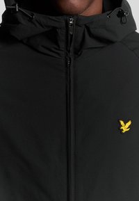 Veste à capuche noire avec une fermeture éclair intégrale, présentant une finition texturée et un logo d'aigle jaune sur la poitrine. Cordon de serrage ajustable au niveau de la capuche.