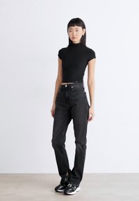 Must mustriba crop-top, kõrge vöökohaga mustad teksad ja mustad tossud. Mudel seisab ühetaolise heleda tausta ääres.