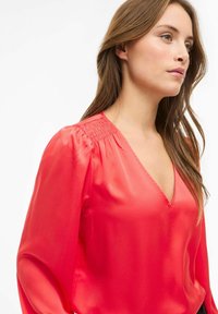 Blusa de satén roja con escote en V, mangas largas y abullonadas, y detalles fruncidos en los hombros. Textura suave con un ligero acabado satinado.