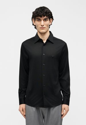 Muž s kučeravými čiernymi vlasmi, oblečený v čiernej košeli na gombíky a sivých nohaviciach, stojaci pred jednoduchým svetlým pozadím.