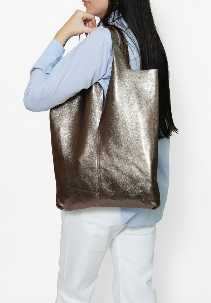 Metalen bronzen tote bag met een gladde textuur, grote rechthoekige vorm en dubbele handvatten, gezien over de schouder van een persoon geslingerd.