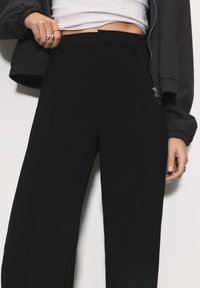 Črne hlače visoke pasu s ravnimi nogavicami, ki imajo urejene plise in teksturirano blago. Okrašene z detajlom srebrnega zadrge.