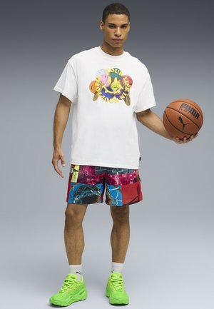 Giovane uomo che tiene un pallone da basket, indossa una maglietta bianca con stampa grafica, pantaloncini da basket colorati, scarpe da ginnastica verde neon, e si trova su uno sfondo grigio.