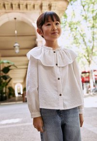 Blouse blanche à boutons avec col à volants, tissu texturé et manches longues, associée à un jean en denim bleu. Cadre extérieur avec verdure.