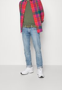 Czerwona i różowa koszula w kratę zapinana na guziki, nałożona na oliwkową koszulkę; jasnoniebieskie dżinsy; białe sneakersy z czarnymi akcentami.