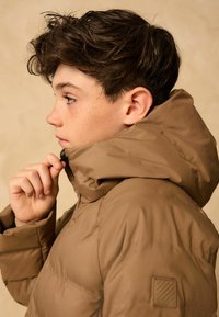 Veste matelassée marron avec un col haut, tissu texturé, et fermeture éclair ; logo sur la manche ; portée par une personne de profil gauche.