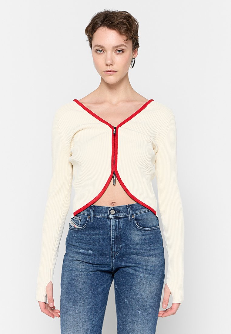 Cardigan corto a maniche lunghe a coste color crema con chiusura a zip rossa e dettagli rossi lungo il collo, abbinato a jeans in denim blu.