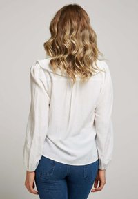 Blouse en coton blanc avec de longues manches bouffantes, un col délicat et un ourlet incurvé. Portée avec un jean bleu ajusté, affichant un design décontracté.