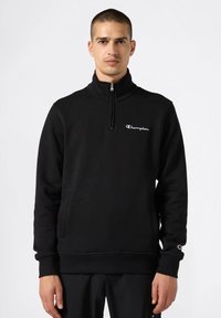 Felpa nera con zip realizzata in materiale morbido, caratterizzata da un collo alto, tasche frontali e un piccolo logo Champion bianco sul petto.