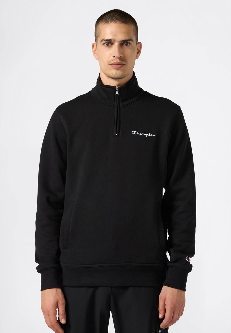 Felpa nera con zip realizzata in materiale morbido, caratterizzata da un collo alto, tasche frontali e un piccolo logo Champion bianco sul petto.