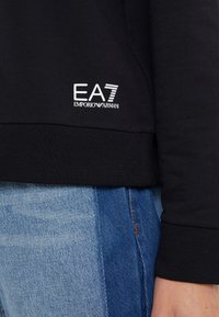 Sudadera negra con ribetes en el dobladillo y los puños, con un logo blanco de EA7 Emporio Armani en el frente. Combinada con jeans azules.