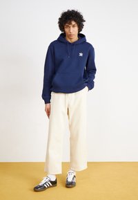Námořnicky modrá mikina s kapucí s bílým logem, krémové široké kalhoty a černo-bílé sportovní boty; hladká látka s volným střihem.