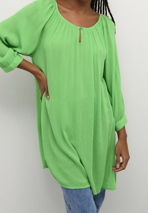 Femme portant un chemisier ample à manches longues de couleur vert vif avec un col froncé et un détail bouton en forme de goutte, les mains posées sur la hanche et le côté.