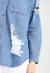 Chemise en denim bleu clair avec trous usés et effilochés sur la manche et le devant, boutons-pression argentés, portée avec un pantalon blanc.