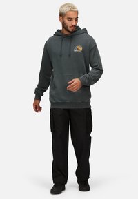 Tmavě šedá mikina s kapucí a přední kapsou, na které je vyšívané logo. V kombinaci s černými cargo kalhotami a černými teniskami.