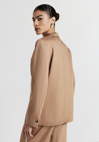 Blazer oversize beige avec une texture lisse et des épaules structurées. Le dos présente une couture centrale unique et des détails de boutons subtils.