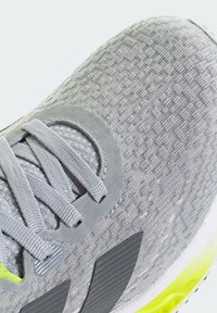 Zapatilla deportiva gris que presenta una parte superior de malla texturizada, cordones grises y un acento amarillo brillante en la suela. Incluye detalle de logo en relieve.