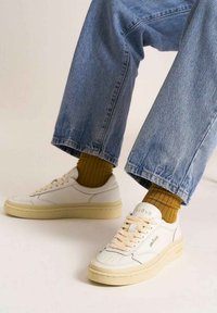 Sneakers bianchi in pelle con suole in gomma beige, caratterizzati da dettagli perforati, punta rotonda e lacci crema. Abbinati a jeans blu e calzini a coste gialli.
