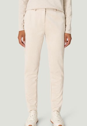 Person trägt beige, eng anliegende Hose, ein beiges, strukturiertes Langarmoberteil und weiße Turnschuhe mit braunen Sohlen, steht vor einem schlichten Hintergrund.