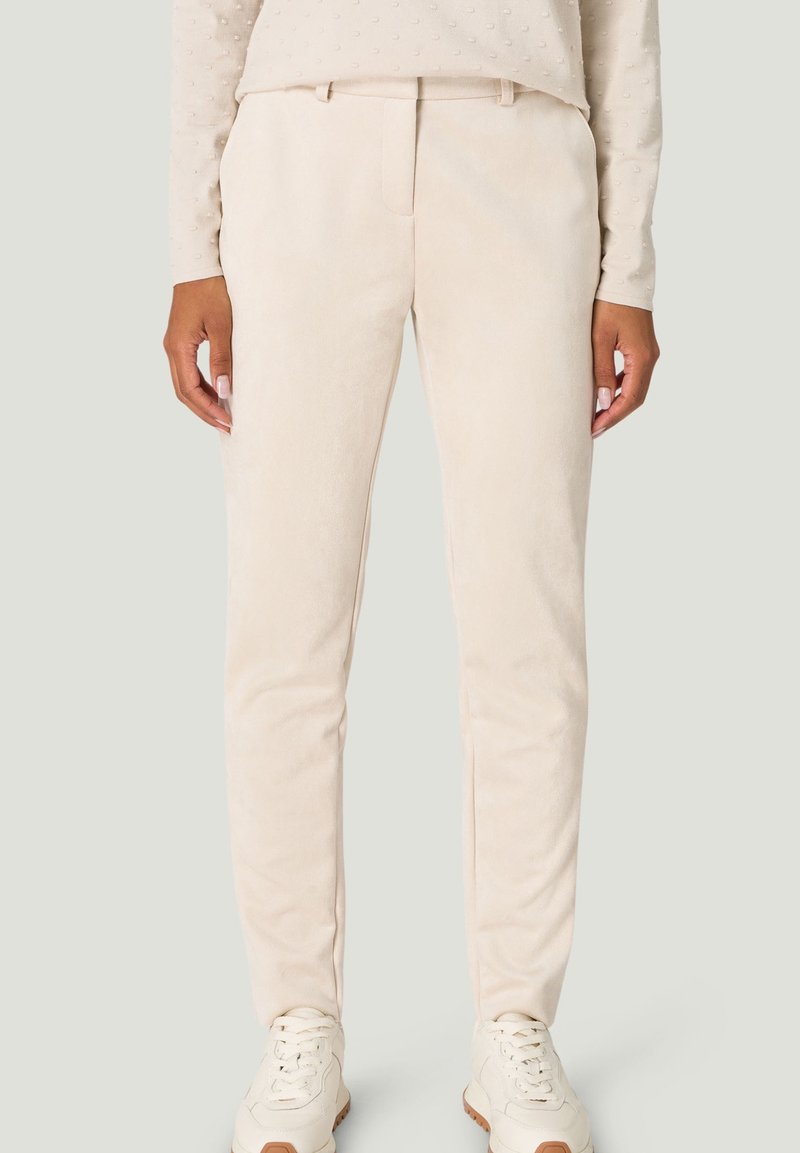 Personne portant un pantalon beige coupe slim, un haut à manches longues texturé beige, et des baskets blanches avec des semelles marron, debout devant un fond uni.