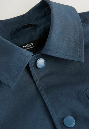 Tumed sinine nööpidega pluus peente vertikaalsete triipudega. Omab teravat kraed, ühte nööpi kinnitust ning pehmet, siledat tekstuuri.