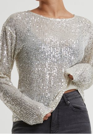 Persoon die een zilverkleurige, met pailletten versierde top met lange mouwen draagt, lichtjes ingestopt in een donkere, hooggetailleerde jeans, met één hand die de zoom rechtzet.