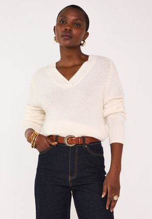 Femme portant un pull blanc en V, un jean foncé avec une ceinture marron, des boucles d'oreilles en or, des bracelets et une grande bague en or, posant sur fond blanc.