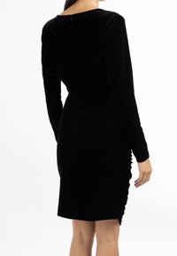 Svart sammetsklänning med långa ärmar, rundad halsringning och rynkad detalj på sidan. Stängs bak med en diskret dragkedja.