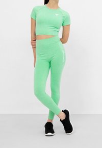 Osoba oblečená v světle zeleném krátkém crop topu a odpovídajících legínách s vysokým pasem, s černými teniskami, stojí se zkříženýma nohama a jednou rukou za zády.