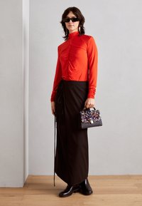 Kvinna som bär svarta solglasögon, röd långärmad pösig topp, lång svart kjol, svarta stövlar, och håller en liten paljettväska.