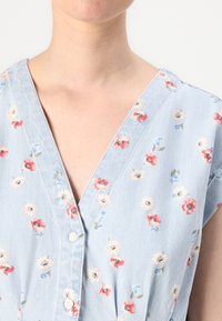 Απαλή μπλε floral φόρεμα με V-λαιμόκοψη, κοντά μανίκια και λεπτομέρειες με κουμπιά. Διαθέτει μικρά μοτίβα κόκκινων, ροζ και λευκών λουλουδιών στο ύφασμα.