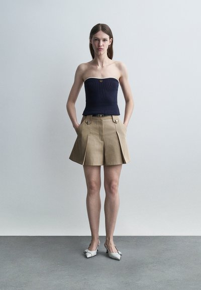 Νεαρή γυναίκα που στέκεται με τα χέρια στις τσέπες, φορώντας αμάνικη μπλε μπλούζα, μπεζ πλισέ σορτς και λευκές μυτερές γόβες, με απλό φόντο.