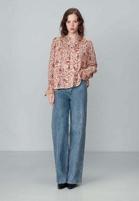 Blouse à manches longues, à boutons, en tissus à motifs rouge, beige et marron, associée à un jean en denim taille haute et à jambe large de couleur bleu clair.
