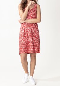Robe rouge sans manches avec un motif floral blanc, tissu texturé et forme évasée, assortie à des baskets blanches.