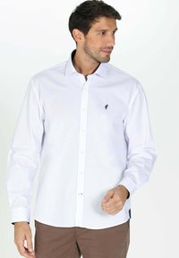 Chemise blanche à manches longues avec un col boutonné, comportant un petit logo noir sur la poitrine. Tissu en coton, coupe normale, texture lisse.