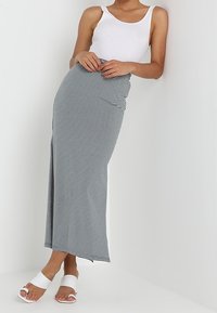 Femme portant une robe maxi ajustée à rayures noires et blanches, un haut sans manches blanc et des sandales blanches ouvertes, se tenant contre un mur blanc.