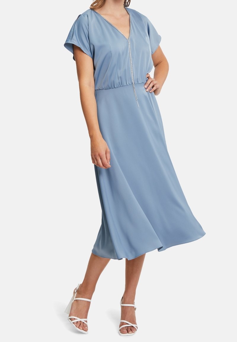 Femme portant une robe midi bleu clair à manches courtes, encolure en V et galon orné de strass, associée à des sandales blanches à talons hauts et à brides.