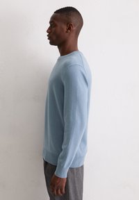 Jasnoniebieski dziergany sweter z okrągłym dekoltem i ściągaczami na rękawach, noszony na szarych spodniach, prezentowany na jednolitym białym tle.