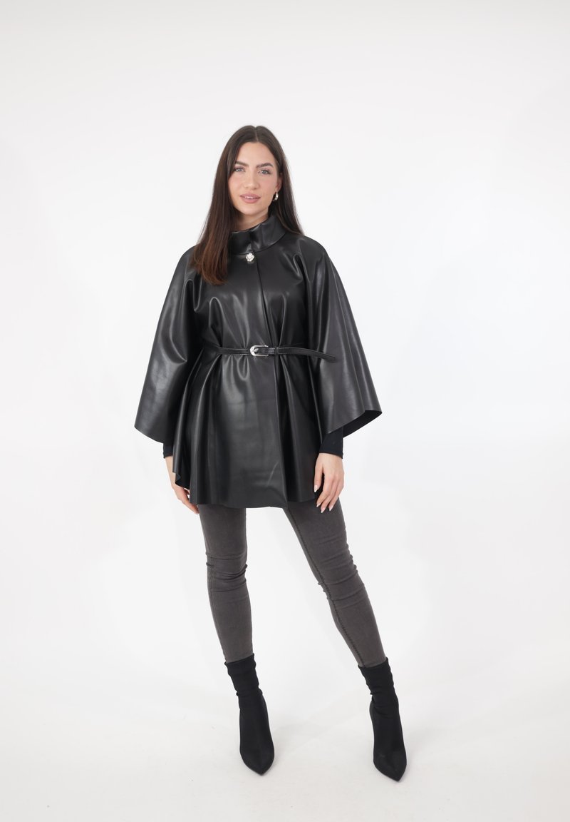 Czarna sukienka w formie peleryny z ekologicznej skóry z wysokim kołnierzem, paskiem podkreślającym talię, rozkloszowanymi rękawami i połyskliwą teksturą; zestawiona z dopasowanymi szarymi jeansami i czarnymi botkami.